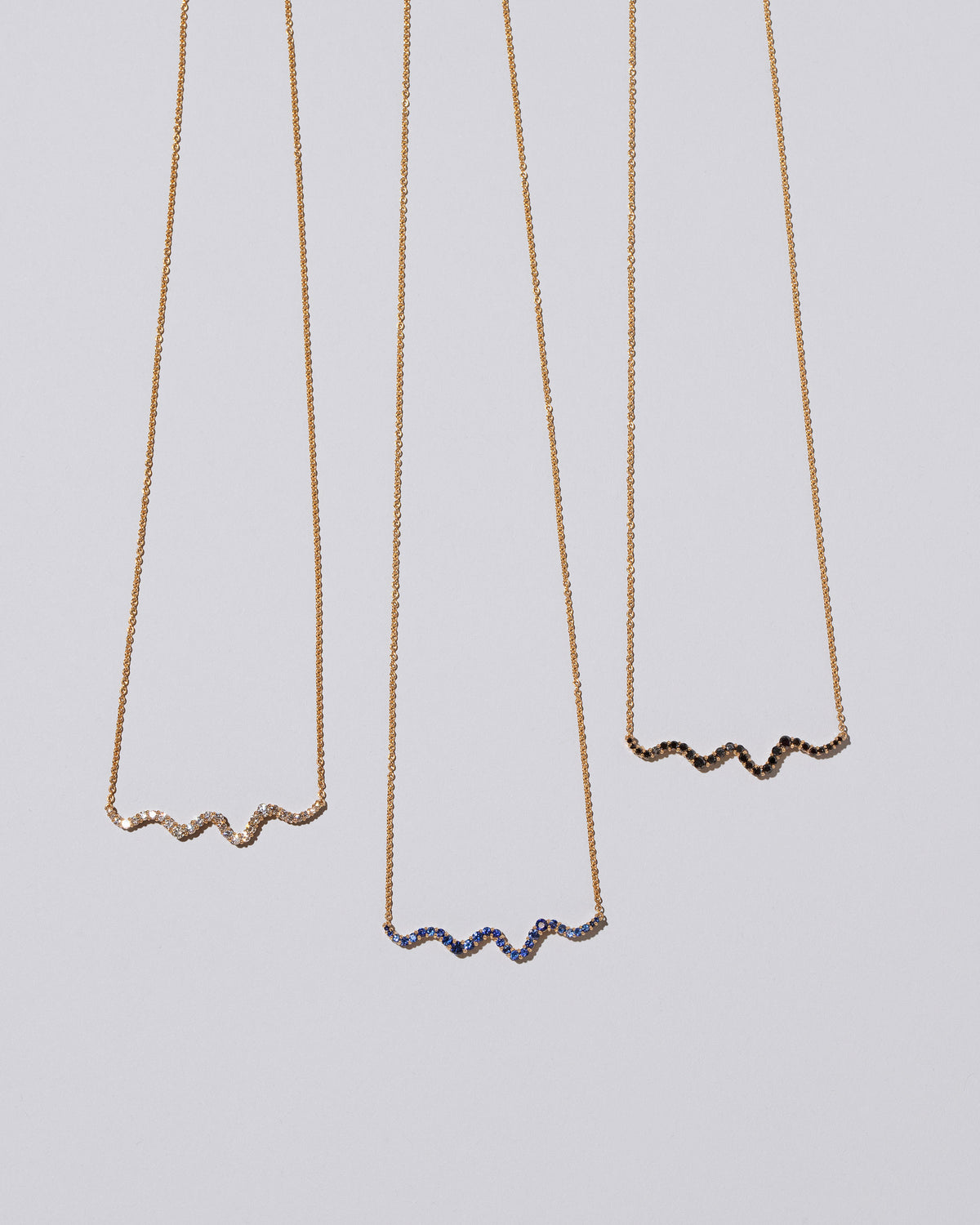 Closeup details of a group of Blue Sapphire, Black Diamond and White Diamond Ripple Wave Necklaces on light color background.